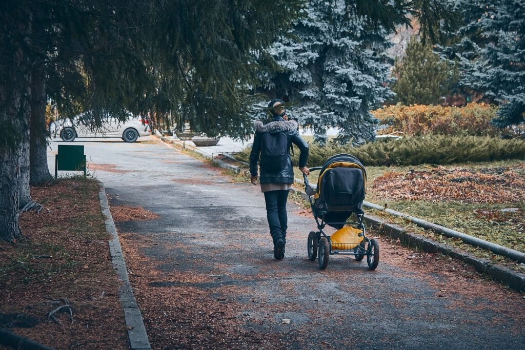 A parent with a backpack pushing a pram along a path through an autumn park